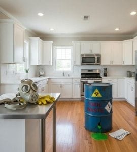 A bright clean kitchen with a drum containing toxic chemicals, a gas mask on the countertop with some yellow thick rubber gloves, a cloth for cleaning and a mop next to the drum of toxic chemicals.